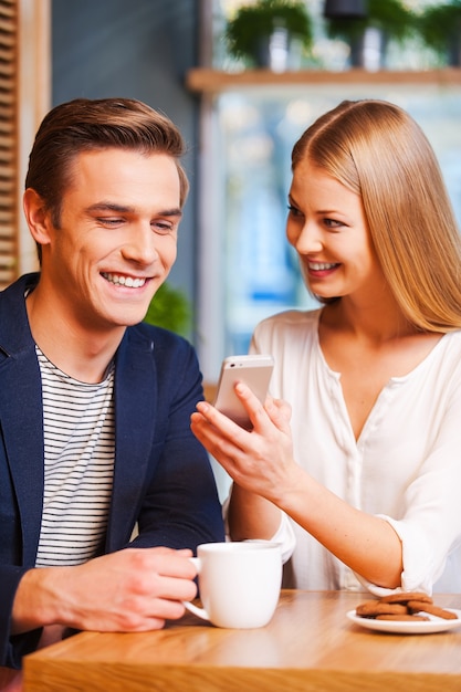 Olha para esta fotografia! Mulher jovem e bonita mostrando algo no celular para o namorado enquanto tomam um café juntos