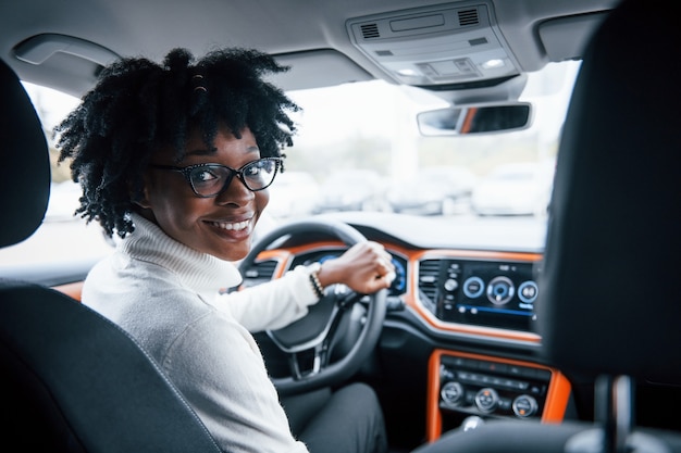 Olha para trás. Jovem mulher afro-americana se senta dentro de um carro novo e moderno.