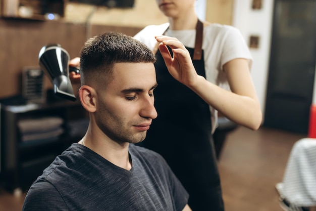 Olhando bem já feche a vista lateral do jovem sendo preparado pelo cabeleireiro com secador de cabelo na barbearia