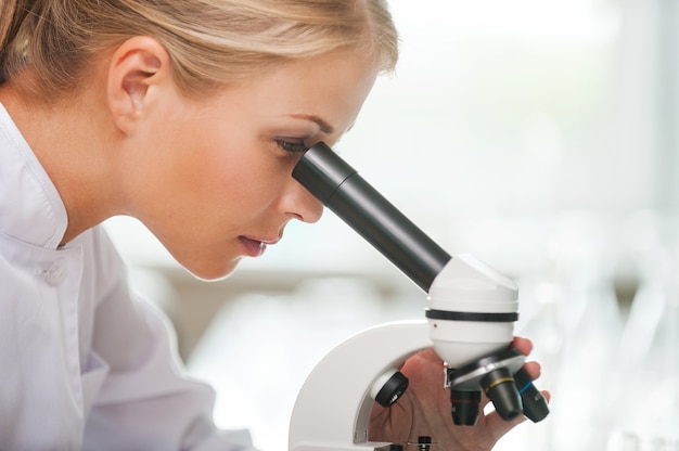 Olhando mais de perto as bactérias. Vista lateral de uma jovem cientista concentrada usando microscópio enquanto trabalhava no laboratório