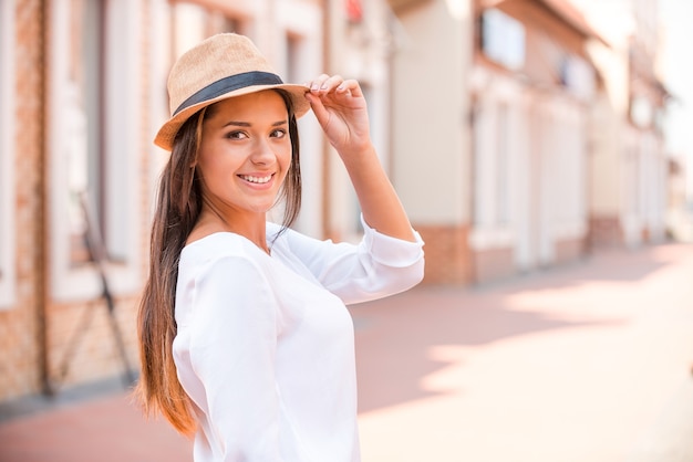 Olhando na moda. Mulher jovem e bonita ajustando o chapéu e sorrindo ao ar livre