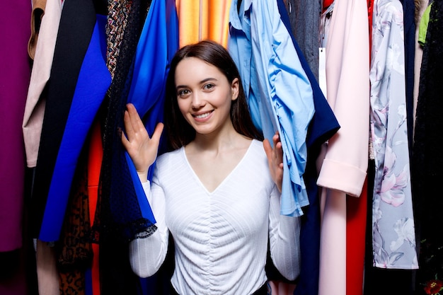 Olhando para a câmera, uma jovem bonita entre as roupas penduradas no cabide afasta as mãos