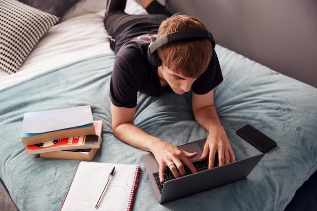 Olhando para baixo no estudante universitário masculino usando fones de ouvido deitado na cama trabalhando no laptop