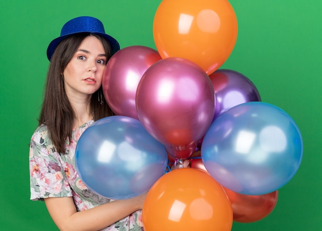 Olhando uma jovem linda com chapéu de festa segurando balões isolados na parede verde