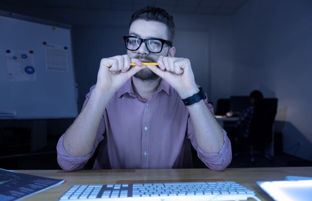 Foto olhar pensativo. homem agradável e barbudo sentado em frente à tela do computador e olhando para ele enquanto segura um lápis