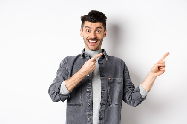 Olhe aqui. Homem elegante feliz sorrindo, apontando os dedos para o espaço vazio, mostrando a oferta promocional legal, em pé na jaqueta jeans em fundo branco.