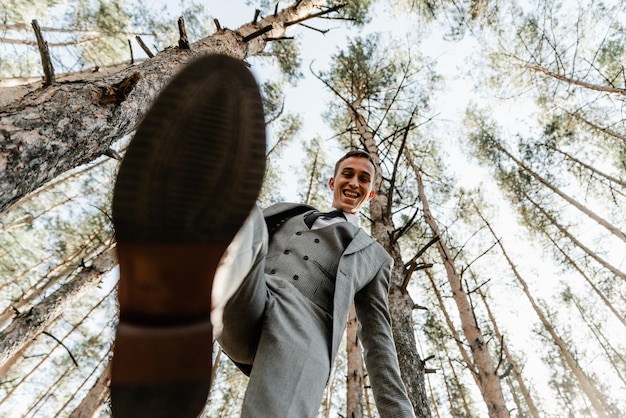 Olhe de baixo para o noivo de terno que está na floresta. noivo com uma perna levantada. um cara de aparência europeia está se divertindo. emoções diferentes de um homem. noivo no dia do casamento