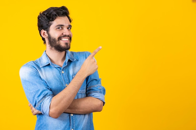 Olhe lá, jovem bonito feliz na camisa jeans apontando para longe e sorrindo em pé contra um fundo amarelo