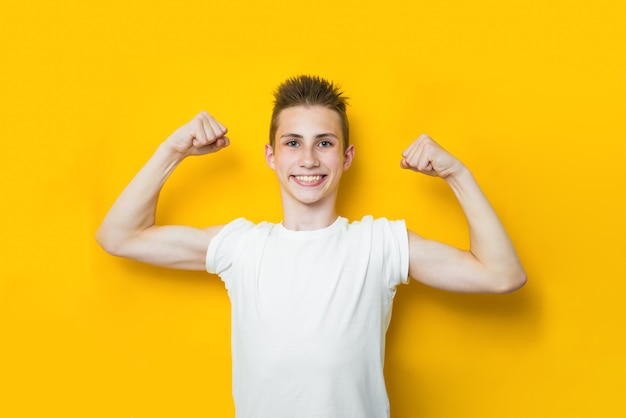 Olhe meu bíceps. Foto de menino adolescente sorridente engraçado em t-shirt branca casual posando isolado em fundo amarelo de cor brilhante. Conceito de infância, fitness e esportes