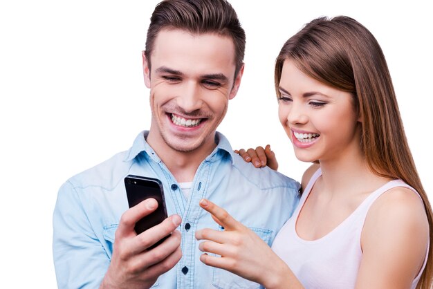 Olhe para essa foto! Jovem casal apaixonado sorrindo e olhando para o telefone celular, enquanto se apoia em um fundo branco