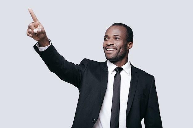 Olhe para lá! Africano jovem bonito em trajes formais apontando para longe e sorrindo em pé contra um fundo cinza