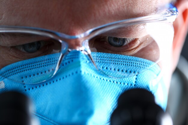 Olhos de trabalhador de laboratório masculino olhando para microscópio usando máscara protetora