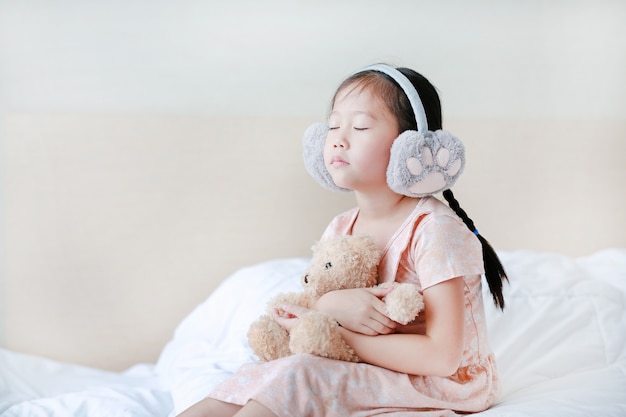Olhos fechados menina vestindo inverno capas para as orelhas e abraçando ursinho enquanto está sentado na cama em casa.