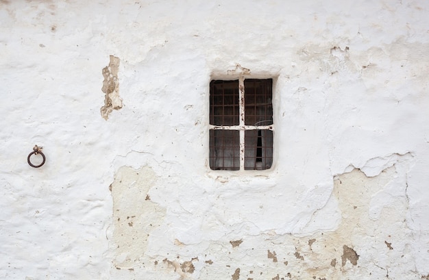 Oliva de Plasencia, España - 8 de abril de 2021: Una ventana vieja con dos rejas de hierro forjado en una pared encalada y junto a un viejo anillo de hierro oxidado para atar a los animales.