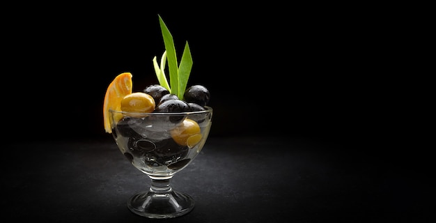 Oliven auf schwarzem Grund, in einer Glasschüssel, mit einer Scheibe Orangen- und Frühlingszwiebelblättern