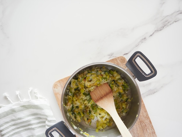 Olla de sopa de verduras con espátula de madera sobre una encimera de cocina de mármol, tablero de madera y paño de cocina