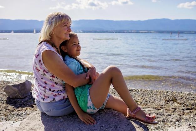 Oma mit Mädchen sitzt am Ufer des Genfer Sees und blickt auf Wasser- und Bergsommerreisen