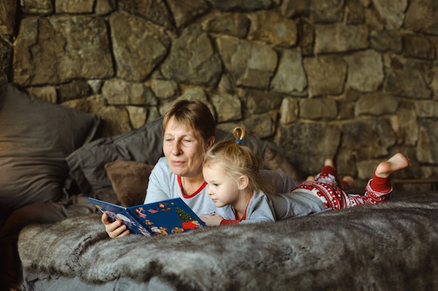 Oma und Enkelin im Weihnachtspyjama lesen ein Buch und liegen auf dem Bett im Chalet. Familienweihnachten.