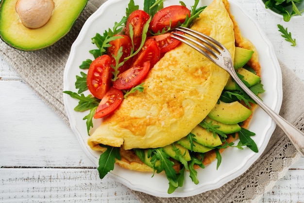 Omelete com abacate, tomate e rúcula em prato de cerâmica branco sobre superfície de pedra clara