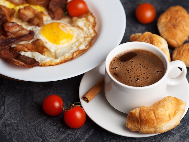 Omelete com fatias de bacon e tomate em um prato branco, uma xícara de café expresso com croissants