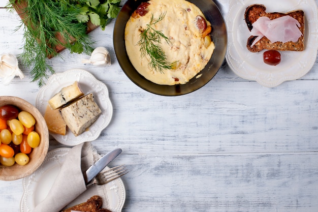 omelete com tomate em uma frigideira no café da manhã, queijo e verduras em um fundo branco de madeira