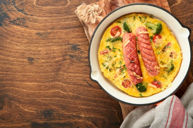 Omelete ou fritada com duas salsichas queijo derretido tomate cereja brócolis e salsa na frigideira vermelha ou panela de ferro sobre fundo de mesa rústica de madeira velha Conceito de comida de café da manhã saudável Vista superior