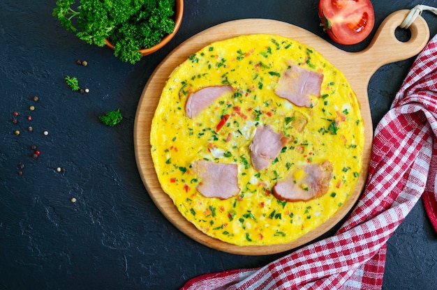 Omelett mit Schinken, Käse, Gemüse auf einem Holzbrett auf einem schwarzen Hintergrund. Fritata - ein köstliches, gesundes Frühstück. Draufsicht, flach liegen.