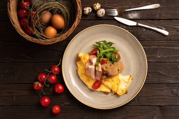 Omelett mit Speck, Kirschtomaten, Rucola und Brot wird wunderschön auf einem dunklen Holztisch serviert.