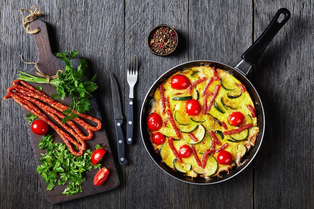 Omelett mit Zucchini, dünnen geräucherten Würsten und Tomaten in einer Bratpfanne auf einem dunklen Holztisch mit Zutaten auf einem Schneidebrett, horizontale Ansicht von oben, flach