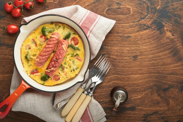 Omelette oder Frittata mit zwei Würstchen geschmolzenem Käse, Kirschtomaten, Brokkoli und Petersilie auf roter Pfanne oder Eisenpfanne auf altem Holztisch im Landhausstil Gesundes Frühstückskonzept Draufsicht