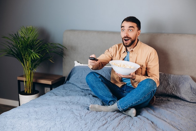 Omg, o que é isso. homem espantado com o tempo livre de quarentena, assistir filme impressionado, surpreso e inesperado filme de suspense terminando segurar uma grande caixa de pipoca sentar sofá dentro de casa