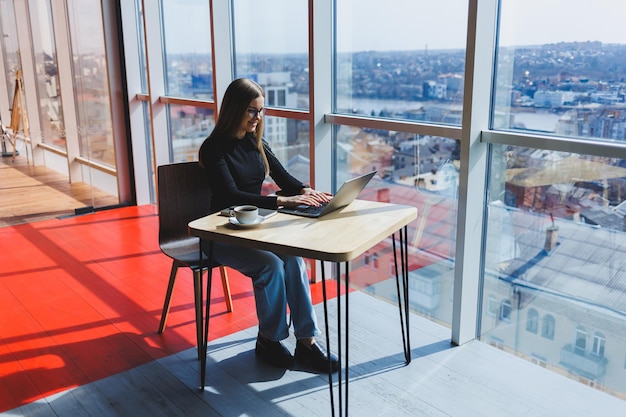 Online-Arbeit in einem Café Fröhliches Mädchen in Freizeitkleidung und Brille mit einem Laptop Fernarbeit oder Bildung mit modernen digitalen Technologien für einen komfortablen Lebensstil in der Stadt