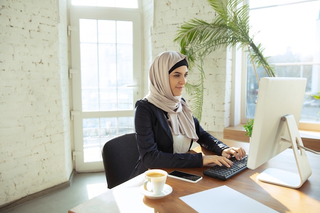 Online arbeiten. Porträt einer schönen arabischen Geschäftsfrau, die Hijab trägt, während sie im Openspace oder im Büro arbeitet. Berufskonzept, Freiheit im Geschäftsbereich, Führung, Erfolg, moderne Lösung.