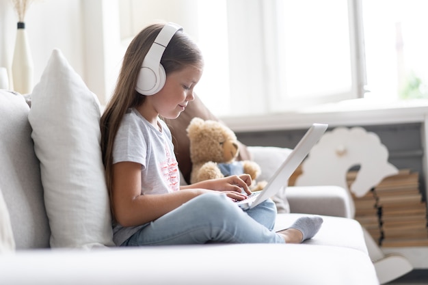 Online-Bildung von Kindern. Süßes Mädchen mit Kopfhörern, das zu Hause auf der Couch sitzt und einen Video-Lehrer-Konferenz-Laptop sieht.