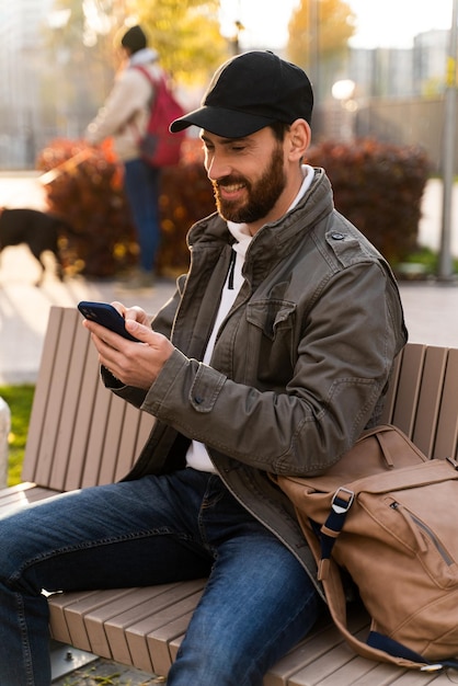 Online Chat. Lächelnder junger Mann, der modernes Smartphone an der Straße verwendet. Hübscher brünette Kerl, der Nachrichten überprüft, während er auf der Bank sitzt. Zeitgenössisches Lebenskonzept