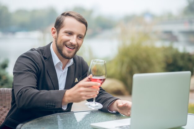 Online-Dating. Junger bärtiger attraktiver Mann mit Glas Wein, der online am Laptop im Sommercafé im Freien plaudert