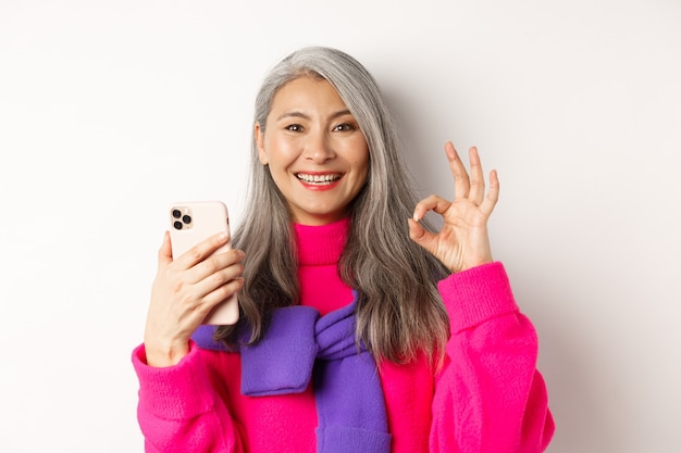 Online Einkaufen. Nahaufnahme einer modischen asiatischen Seniorin, die ein okayzeichen zeigt, Smartphone verwendet und etwas genehmigt, stehend auf weißem Hintergrund