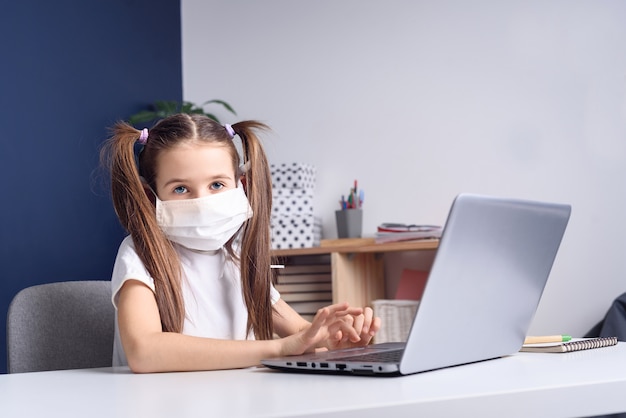 Online-Fernunterricht. Schulmädchen in der medizinischen Maske, die zu Hause studiert, am Laptop-Notizbuch arbeitet und Hausaufgaben macht. Coronavirus-Quarantänekonzept