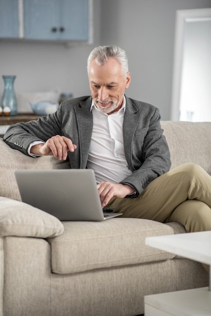 Online-Job. Erfreute ältere männliche Person, die Positivität ausdrückt, während sie Nachrichten prüft und auf seinem Sofa sitzt