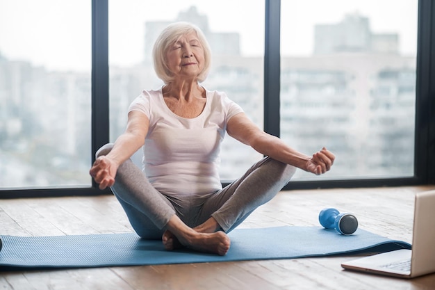 Online-Klasse. Grauhaarige Seniorin, die auf dem Boden sitzt und einen Online-Yoga-Kurs hat