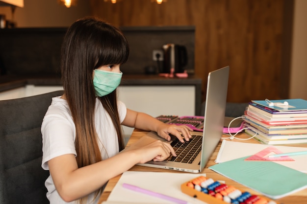 Online-Lernen während der Quarantäne. Mädchen mit einer Schutzmaske auf ihrem Gesicht macht Hausaufgaben zu Hause.