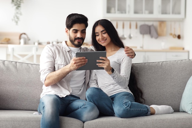 Online-Meeting mit Familie, Konferenz, soziale Distanzierung und neue Normalität, Covid-19. Lächelnde tausendjährige arabische Frau und Ehemann schauen auf Tablet und chatten mit Freunden, auf der Couch im Wohnzimmer?