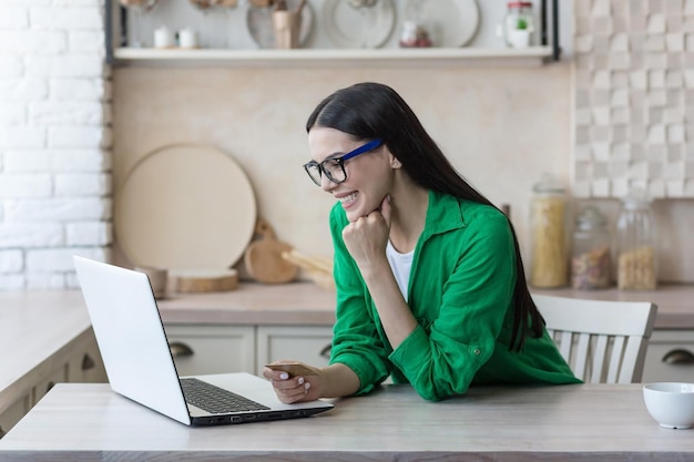 Online-Shopping Eine junge schöne Frau mit Brille und grünem Hemd sitzt zu Hause mit einem Laptop