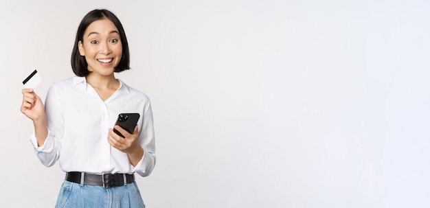 Online-Shopping-Konzept Bild einer jungen asiatischen modernen Frau mit Kreditkarte und Smartphone-Kauf mit Smartphone-App, die kontaktlos auf weißem Hintergrund bezahlt