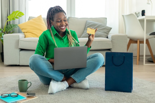 Online-Shopping-Konzept Fröhliche schwarze Frau, die Kreditkarte zeigt und Laptop-Kleidung über das Internet bestellt