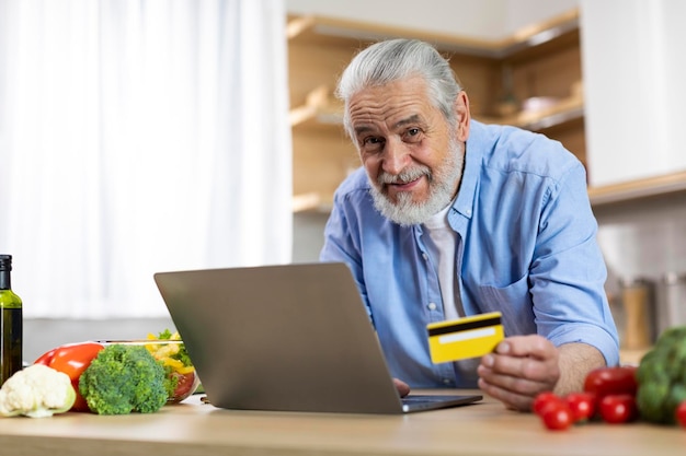 Online-Shopping Lächelnder älterer Mann mit Laptop und Kreditkarte in der Küche
