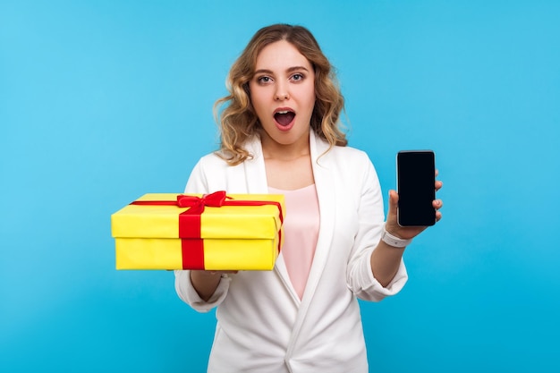 Online-Shopping Porträt einer erstaunten schönen Frau mit welligem Haar in weißer Jacke, die eine Geschenkbox und ein Telefon hält und überrascht auf die Kamera blickt, die im Innenbereich auf blauem Hintergrund aufgenommen wurde