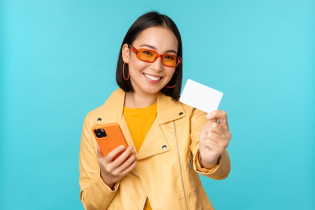 Online-Shopping Stilvolle junge asiatische Frau mit Sonnenbrille, die Kreditkarte zeigt und mit dem Smartphone im Internet bezahlt und einen Kauf über blauem Hintergrund tätigt