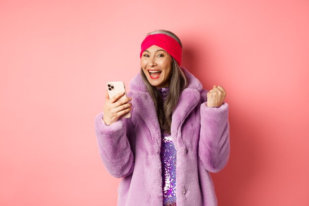 Online-Shopping und Modekonzept. Fröhliche asiatische Seniorin, die den Preis im Internet gewinnt, das Handy hält und die Faustpumpe macht, den Freudenschrei, der über rosafarbenem Hintergrund steht.