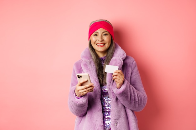 Online-Shopping und Modekonzept. Lächelnde Frau mittleren Alters im stilvollen Pelzmantel unter Verwendung des Mobiltelefons und der Plastikkreditkarte, die glücklich auf Rosa stehen.